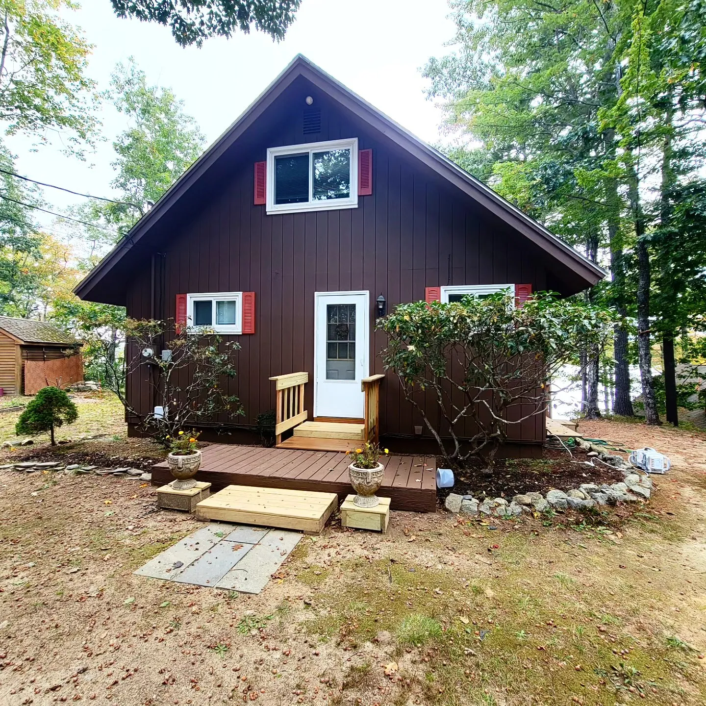 A dark-stained wood cottage with red shutters, fresh deck boards and trimmed shrubs, the finish new and the trim crisp